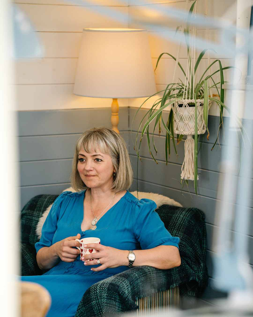 Woman in a blue dress sitting on a couch holding a cup, with a lamp and plant in the background.